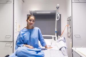 Young female patient in hospital ward,dressed in blue gown,sitting on bed and waiting to be examined by doctor,smiling softly and satisfied with service and services provided,individual hospital room photo
