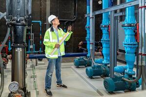 asian man worker in protective uniform and with hardhat using tablet for checking temperature in pipes Factory interior. photo