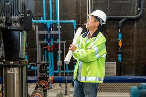 asian man worker in protective uniform and with hardhat using tablet for checking temperature in pipes Factory interior. photo