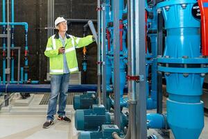 asian man worker in protective uniform and with hardhat using tablet for checking temperature in pipes Factory interior. photo
