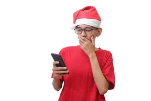Portrait of excited Asian man in red t-shirt with santa hat looking at his mobile phone while covering his mouth with fingers. Wow face expression. Isolated image on white background photo