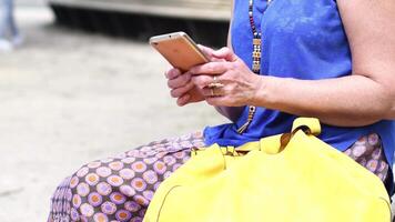 unrecognizable middle-aged woman in a blue blouse and a yellow bag is typing a message on a smartphone with two fingers,sitting on a park bench,using messenger in applications,High quality 4k footage video