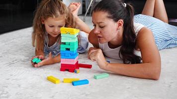 Little girl with her mother playing Jenga board game at home, taking colorful blocks from wooden tower. High quality 4k footage video