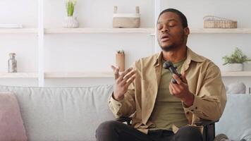A man sits on a couch in his living room. He is wearing a beige button-down shirt over a green t-shirt. He holds a game controller in his hands and looks at the camera. video