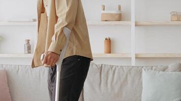 A young man stands in his living room, using crutches for support. He is adjusting his shirt with one hand while balancing with the other. The atmosphere is casual and relaxed. video