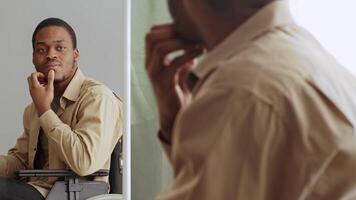 A man seated in a wheelchair examines himself in a mirror, grooming with a thoughtful expression. The setting suggests a personal moment of self-care in a bright environment. video
