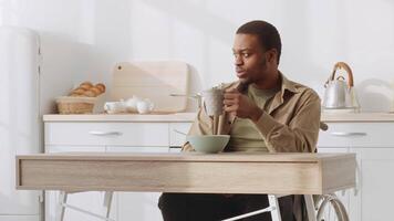 African American man sits at a modern kitchen table, sipping coffee from a mug while gazing thoughtfully out of a window, with fresh pastries in the background and light streaming in. video