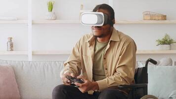 African American man sits in a wheelchair wearing a virtual reality headset and holding a game controller in front of a couch in his living room. video