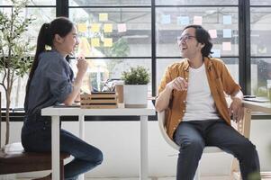 Two Asian workers sitting in chairs at working desks, collaborating and talking at daylight windows in casual small business office, online business job coworkers, positive relationship colleagues. photo