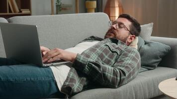 Lazy relaxing lying on sofa couch Caucasian man male freelancer businessman business guy online shopping order food delivery laptop computer working pc browsing net playing internet game web at home video