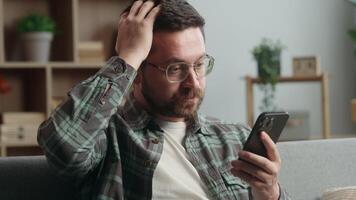 Caucasian man male guy user at home browsing mobile phone app social media smartphone shock bad news amazed trouble problem confused puzzled wonder online internet web at home couch sofa apartment video