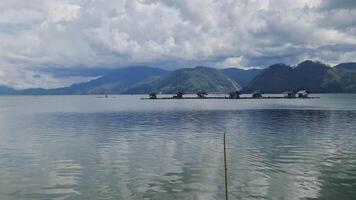 A serene lake with calm waters reflecting the cloudy sky, surrounded by lush green mountains. Small houses on stilts are visible along the shoreline, adding a peaceful, rural charm. video