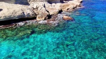 Turquoise waves gently lapping against white rocks create a serene coastal scene. Sunlight shimmering on the water enhances the tranquil beauty of this picturesque landscape video