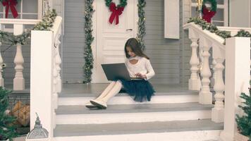 Child making a call using a laptop. The girl sits on the doorstep of her house and communicates via link holding a laptop on her lap video