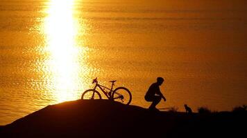 Man with bicycle resting on beach. Silhouette of man resting after cycling trip near river video