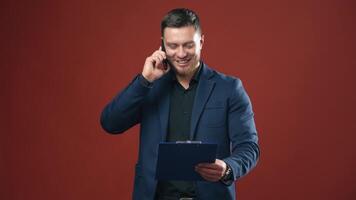 Happy businessman talking the phone. Handsome man in suit holding folder and speaking on the phone in studio. Office manager on dark red background. video