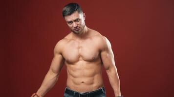 Muscular bodybuilder on dark red background. Athlete with naked torso lifting sausage in one hand and dumbbell in other. Smiling sportsman in studio. video