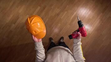 pov first person view Construction worker holding a yellow safety helmet and a red cordless drill, preparing for work on a wooden floor, view from above video