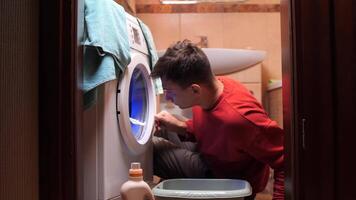 Man watching washing machine carefully. Man looking inside the washing machine. Young man is retrieving laundry from a front-loading washing machine in a dimly lit bathroom, using a small basin video