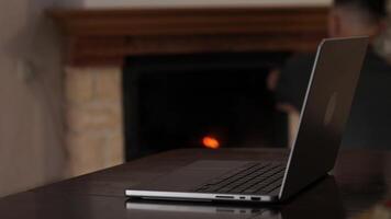 Man sitting at a table works on his laptop, with a lit fireplace creating a cozy atmosphere in the background. The scene captures a comfortable home office setting video