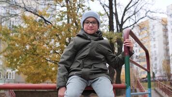 Preteen boy wearing a warm jacket, beanie and glasses is sitting on a climbing frame in a playground during a cloudy autumn day video