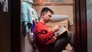 Man in a red shirt reads while waiting for laundry in a cozy bathroom, enjoying a moment of quiet multitasking at home video