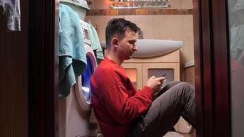 Young man playing game waiting for washing machine at laundry room. man sits on floor near washing machine and uses phone while washing clothes in washing machine video