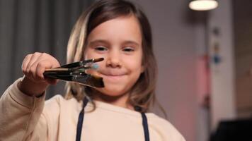 Creative cute girl pose at camera . Young beautiful child looking at camera while standing at room and holding paintbrush. Creative activity. Erudition. child in apron ready for drawing lesson video