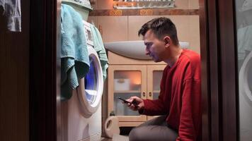 Man is kneeling in the bathroom, using a smartphone while waiting for the washing machine to finish its cycle, with towels hanging on the side. texting video