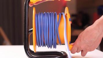 An electrician winds an extension cord onto a plastic and metal reel placed on a white table, showcasing the process of managing electrical tools in a workshop setting video