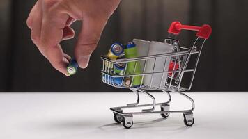 Hand adding a battery to a miniature shopping cart filled with various batteries on a clean white surface showcases consumer behavior in the energy and electronics market video