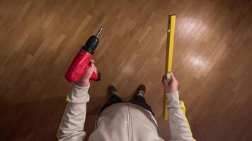 Pov first person view Construction worker holding a level and a cordless screwdriver, standing on a wooden parquet floor, getting ready for work. Man ready to do apartment renovation video