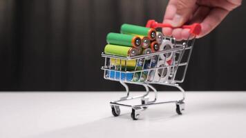 Miniature shopping cart full of colorful cylindrical alkaline batteries is pushed by a hand. The cart is on a white surface with a dark background. concept of buying batteries. video