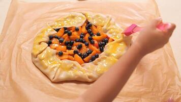 Preparing a fruit galette with care, brushing the crust with egg wash. Close-up of hands adding golden sheen to the edges before oven time. Blueberries and peaches fill the rustic pastry video