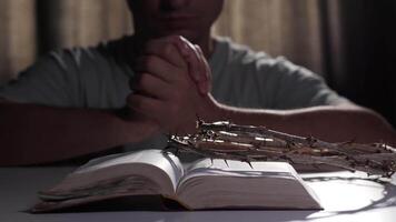 Religious man prays with clasped hands, while a crown of thorns rests on a bible placed on a table, creating a powerful symbol of faith and devotion in a serene indoor setting video