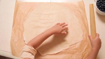 Child using a rolling pin to roll out dough on parchment paper, preparing for baking. Kitchen setup with fresh blueberries in bowls around the table video