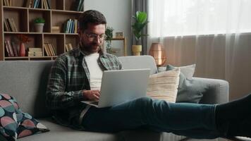 Happy smiling positive Caucasian man male guy at home couch sofa with laptop computer working online internet work typing shopping complete accomplishment done job relax break put hands behind head video
