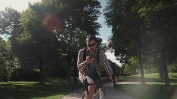 A male with a backpack on a bicycle ride in the park on his cycle talking on the phone in sunny weather. A bicyclist rides his bike in the park and talks on the telephone with a rucksack. video