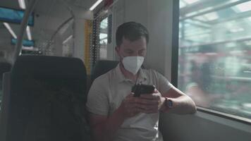 Masked man travels on train by window on summer day in sunny weather and uses smartphone. Man traveler in a mask rides a commuter suburban railway in Germany while surfing the Internet on his phone. video