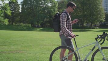 A cyclist walking near his bicycle is chatting online in the park in sunny weather. A commuter with a bicycle and a smartphone walking around. Man enjoys the phone on a summer walk with a bicycle. video