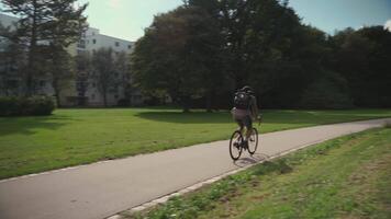 Handsome hipster with backpack and shirt enjoys a bicycle ride through a city park in summer sunny weather. A man riding a city cycle in Germany on a bicycle road. A guy on gravel bicycle in Munich. video
