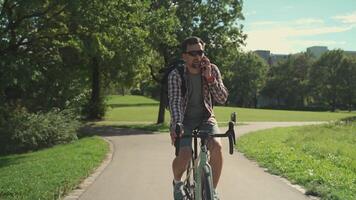 Male cyclist with backpack rides bicycle path in park in summer and talks on his mobile phone. Active lifestyle and commuting on bicycle. Cyclist talking on telephone while cycling in the city park video