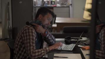 Cervical neck ache, nerve arthritis. Man studying late through night at laptop computer with cervical pain. Exhausted guy relaxing his neck using gun massager while overworking at notebook computer. video