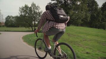 Man in shirt and with rucksack rides bicycle on cycle path in city garden in Munich, Germany. Student cyclist cycling in bikeway in park. A male with a backpack riding a city cycle in a green area. video