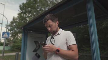 Young caucasian man face beard at the bus stop waiting for the bus in the hands of a mobile phone sends messages. Theme of urban transport and mobility, traveling around Europe in comfortable transport. video