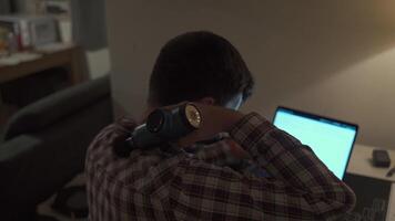 Tired male student studying on laptop at home until late and has neck pain and massages himself with percussion gun. Neck and back pain. Overworked man uses pistol massager to relieve muscle tension video