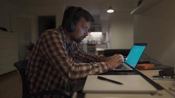 Focused man in headphones with a laptop late at night at home, taking notes, student learning a language, watching an online webinar, hearing an audiocourse, the concept of e-education. Homework. video