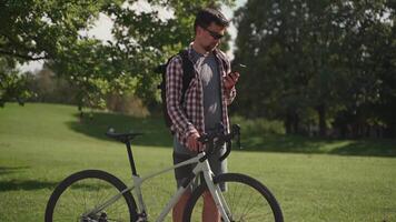 A cyclist walking near his bicycle is chatting online in the park in sunny weather. A commuter with a bicycle and a smartphone walking around. Man enjoys the phone on a summer walk with a bicycle. video