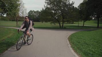 Handsome hipster with backpack and shirt enjoys a bicycle ride through a city park in summer sunny weather. A man riding a city cycle in Germany on a bicycle road. A guy on gravel bicycle in Munich. video