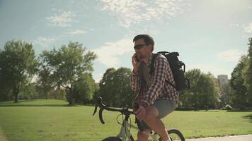 Male cyclist with backpack rides bicycle path in park in summer and talks on his mobile phone. Active lifestyle and commuting on bicycle. Cyclist talking on telephone while cycling in the city park video